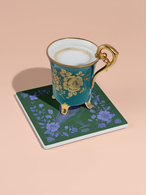 Square ceramic coaster featuring intricate lavender illustration of a raven surrounded by botanical elements on forest green background, with ornate border design and cork backing.